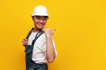 asian male construction worker in uniform and safety glasses pointing back on yellow isolated background