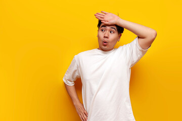 tired young asian guy in white t-shirt wiping sweat from forehead on yellow isolated background