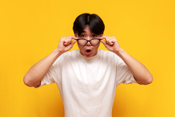 shocked young asian guy in white t-shirt with glasses looking at camera in surprise on yellow isolated background - obrazy, fototapety, plakaty