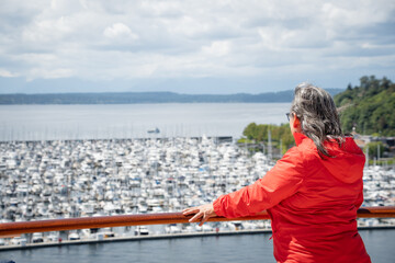 Fototapeta na wymiar Woman with her back to the camera, wearing an orange windbreaker, gazes at the horizon from a marina.