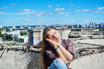 A girl who is not afraid of heights, with a calm face, sits next to a precipice against the backdrop of the city and the blue cloudy sky.