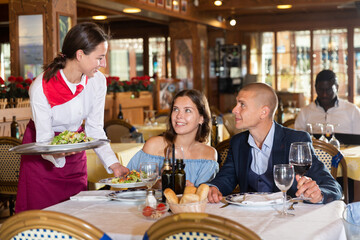 Young attractive waitress brings an order to couple of smiling visitors in a restaurant - obrazy, fototapety, plakaty