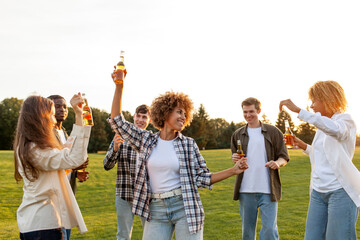 group of young multiracial students holding bottles of beer and dancing at outdoor party,...