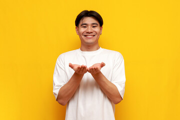 young asian guy in white t-shirt begging and holding empty hands in front of him on yellow isolated background