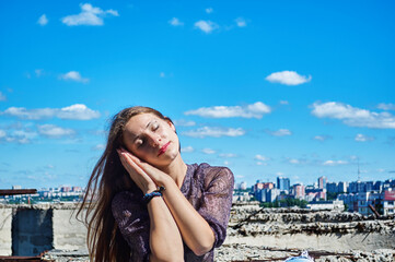 A girl who is not afraid of heights, with a calm face, sits next to a precipice against the backdrop of the city and the blue cloudy sky.