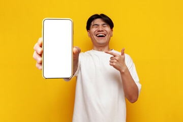 young asian guy in glasses shows blank screen of smartphone and laughs on yellow isolated background - obrazy, fototapety, plakaty