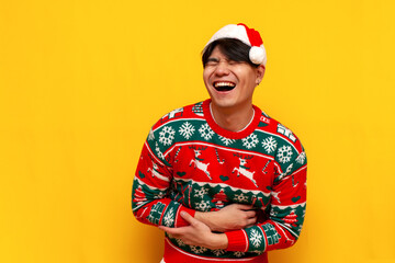 young asian man in christmas sweater and santa hat laughs and makes fun of on yellow isolated...