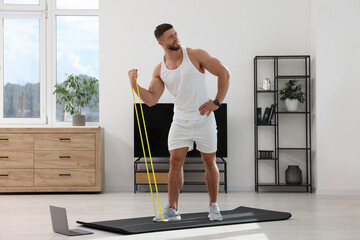Fototapeta premium Muscular man doing exercise with elastic resistance band near laptop on mat at home