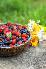 Wicker bowl with different fresh ripe berries and beautiful flowers on wooden surface outdoors