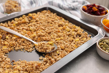 Making granola. Baking tray with mixture of oat flakes, other ingredients and spoon on grey marble table