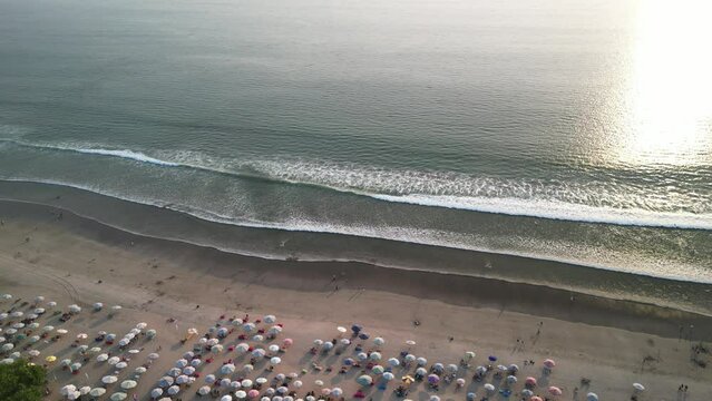 Aerial view of Beautiful sunset on the double six beach with Hotel, resort, cafe buildings, beach umbrella and calm ocean wave in Bali. Tourists are surfing, swimming and walking along the beach.