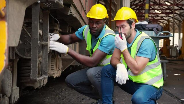 Young caucasian and black engineer maintenance and repair train diesel engine while using talkie with command to worker in station, team engineer inspect system transport, transportation and industry.
