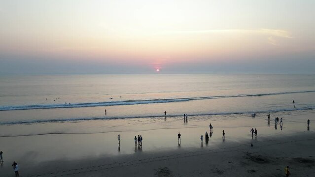 Aerial view of Tourists are enjoying the beautiful sunset on the beach with calm ocean wave in Bali. Tourists are surfing, swimming and walking on the beach at dusk with clear sky.