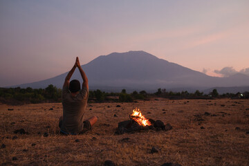 A man does yoga in nature near a fire, practicing alone, against the backdrop of the silhouette of...