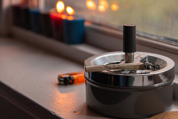 Concept image of a joint (rolled cannabis cigarette) and ashtray placed by the window of a room. Concept of marijuana as a luxury item, social issues, and legalization of cannabis. - obrazy, fototapety, plakaty