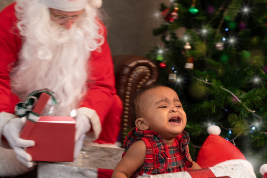 Baby African girl crying, she doesn't like or fear Santa,  Santa comes to play and hold gift box in background, Christmas time together