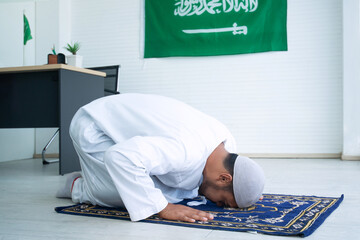 Side view of Arab Muslim man doing Salat on prayer mat at office, Saudi Arabia Flag hanging on wall...