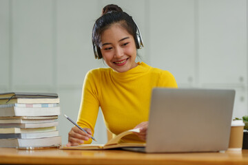 Happy Asian female student wearing wireless headphones studying online, listening to music,...