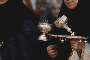 Shallow depth of field (selective focus) details with the hand of a orthodox Christian nun holding a smoking metallic censer incense burner. - obrazy, fototapety, plakaty