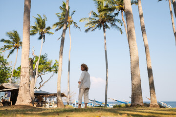 A young girl with blond, curly hair poses in the middle of a palm grove, in beautiful light clothes, on the ocean shore.