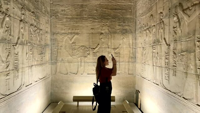 Tourist girl taking photos with her mobile phone to hieroglyphs inside temple of Horus in Edfu, Egypt. Traveler in Egyptian Temple of god Horus in Edfu. Woman in walls with herogliphs. Ancient Egypt.