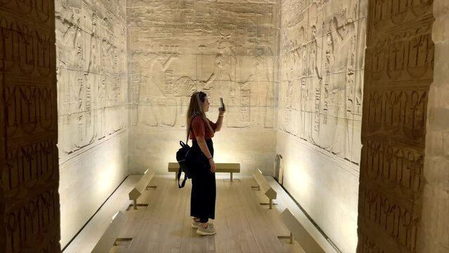 Tourist girl recording videos with her mobile phone to hieroglyphs inside temple of Horus in Edfu, Egypt. Traveler in Egyptian Temple of god Horus in Edfu. Woman in room with herogliphs. Ancient Egypt