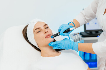 Young woman receiving facial treatment in a professional beauty salon. A close-up cosmetologist manipulates a hydropeeling machine to clean and rejuvenate a patient's skin in a modern clinic.