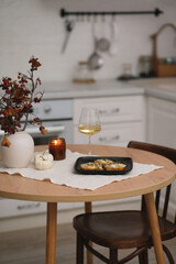 Autumn home interior in kitchen. Light dinner - baked pumpkin with cheese, glass of white wine, burning candle, vase with dry branches on round wooden table in light kitchen.