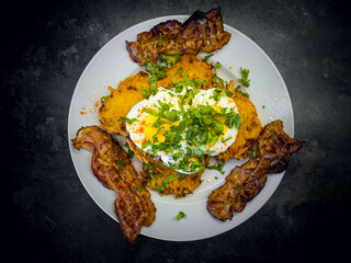 Fried bacon and boiled eggs on a white plate with dark background. Breakfast with bacon, eggs with sour cream topping and lots of parsley on top.