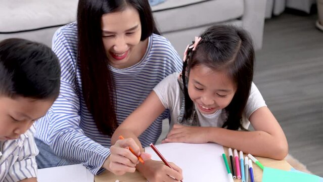 Multitasking mother and teacher teaching homework for cute sibling children, Asian mom take care kids enjoy playtime drawing pictures sitting on the floor together at home in weekend, happy motherhood