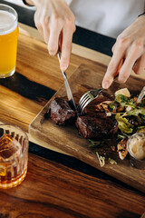 Close up of hands holding knife and fork is cutting grilled ribeye steak on wooden cutting board
