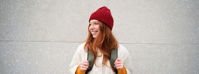 Happy young girl, redhead tourist in red hat with backpack, walks around town, explores city,...