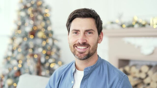 Close up portrait of handsome man sitting in living room at home during winter New Year Xmas holidays. Head shot of a smiling positive bearded male on background of a Christmas tree looking at camera
