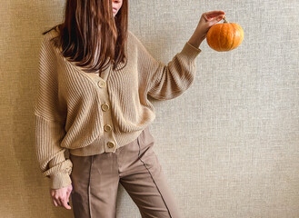 An unrecognizable brunette girl in a beige sweater holds a bright orange pumpkin in her hands on a...