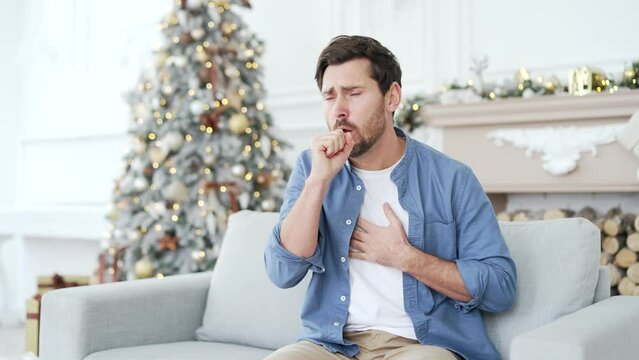 Sick man with cold or flu suffering from cough sitting on sofa at home during winter New Year Christmas Xmas holidays. Upset male feels bad, holds his hand on his chest. He has pneumonia or bronchitis