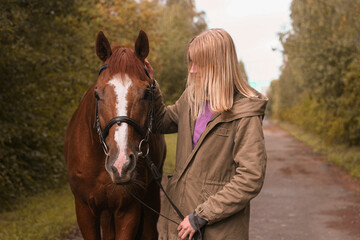 Girl with blond hair in khaki jacket taking care of a red horse in an autumn park