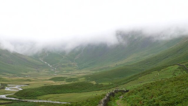 Panning shot of Mosedale Beck stream in the misty mountain valley under low cloud in summer in English Lake District National Park, United Kingdom.