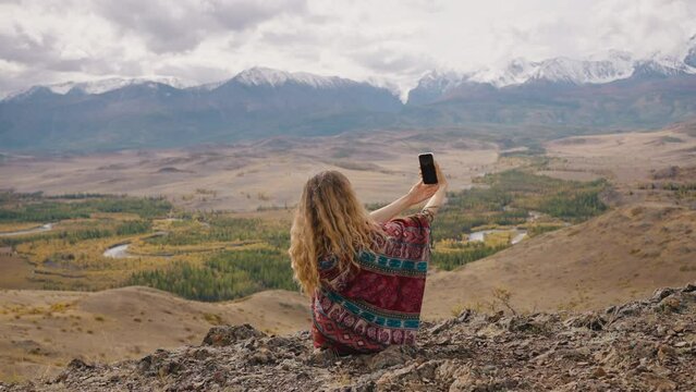 A woman traveler takes a selfie on a smartphone against the backdrop of snowy mountains while sitting on a hill.