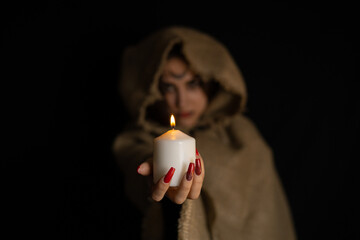 On Halloween night, a hooded witch holds out a burning candle from the darkness. The witch conjures in the evening on halloween over the flame of a candle. Halloween holiday concept.
