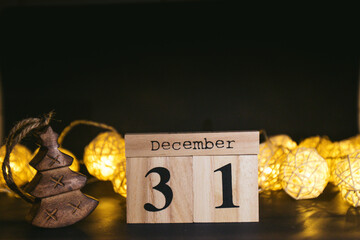 Brown wooden calendar on the black table with New Years garland. Black background. Items to tell date, symbol of appointment, reminder, planner or meeting. Vintage idea shows 31 December, end of year - obrazy, fototapety, plakaty