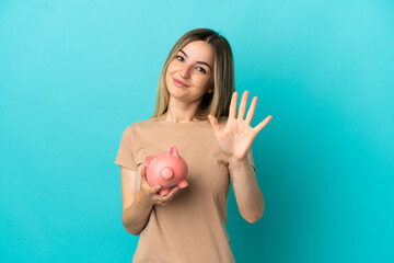 Young woman holding a piggybank over isolated blue background counting five with fingers