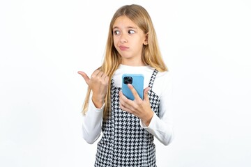 Beautifu blonde kid girl points thumb away and shows blank space aside, holds mobile phone for sending text messages.