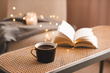 Mug of tea with open paper book on rattan coffee table in bedroom indoors close up over scented...