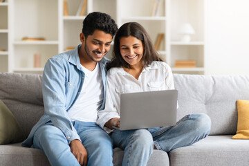 Indian young couple browsing on a laptop, seated on sofa at home