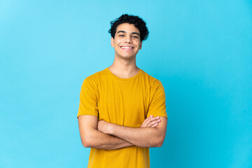 Young Venezuelan man isolated on blue background keeping the arms crossed in frontal position - obrazy, fototapety, plakaty