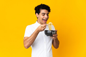 Young Venezuelan man isolated on yellow background holding a bowl of noodles with chopsticks and...