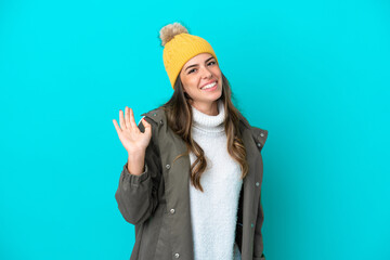 Young Italian woman wearing winter jacket and hat isolated on blue background saluting with hand with happy expression
