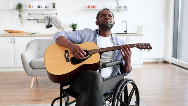 Handsome bearded adult in wheelchair posing with six-string guitar while enjoying pleasant pastime at home. Smiling african man with disability in spectacles learning to play instrument using.