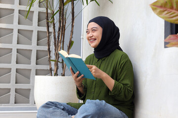 Portrait of attractive Asian hijab woman reading book in balcony. Muslim girl enjoying free time....