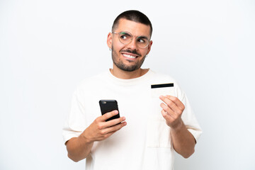 Young caucasian man isolated on white background buying with the mobile with a credit card while thinking